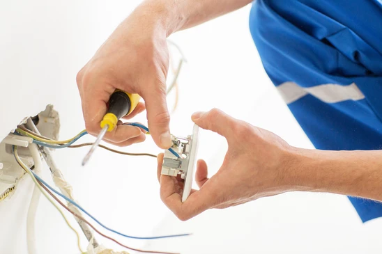 Électricien en bleu travaillant sur installation électrique murale, manipulant câbles et composants avec tournevis