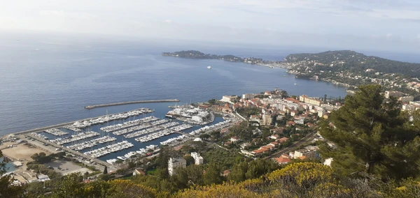 Photographie réaliste montrant une façade de maison ou petit immeuble résidentiel à Beaulieu-sur-Mer ou environnement côtier similaire des Alpes-Maritimes, avec détails architecturaux typiques de la région (architecture méditerranéenne, volets, façade claire). Au premier plan, un électricien en tenue professionnelle (gilet de travail, équipement) effectuant une intervention à un tableau électrique, un coffret ou une installation électrique visible sur la façade ou à l'entrée du bâtiment. Lumière naturelle de jour, style photographie professionnelle de prestation de service.